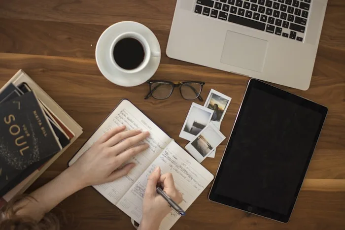 A person writes in a journal with a laptop, book and coffee during creative writing training at CAQA Creative.
