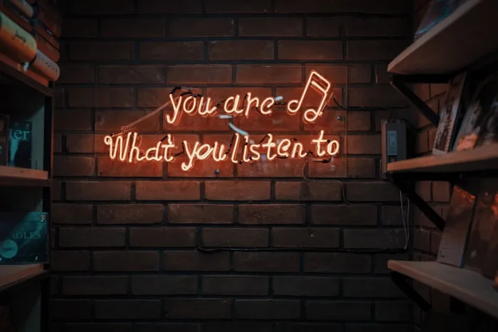 Musical Quote Wall | CAQA Creative A brick wall showcases a neon sign with the quote 'you are What you listen to' and shelves displaying vinyl records.